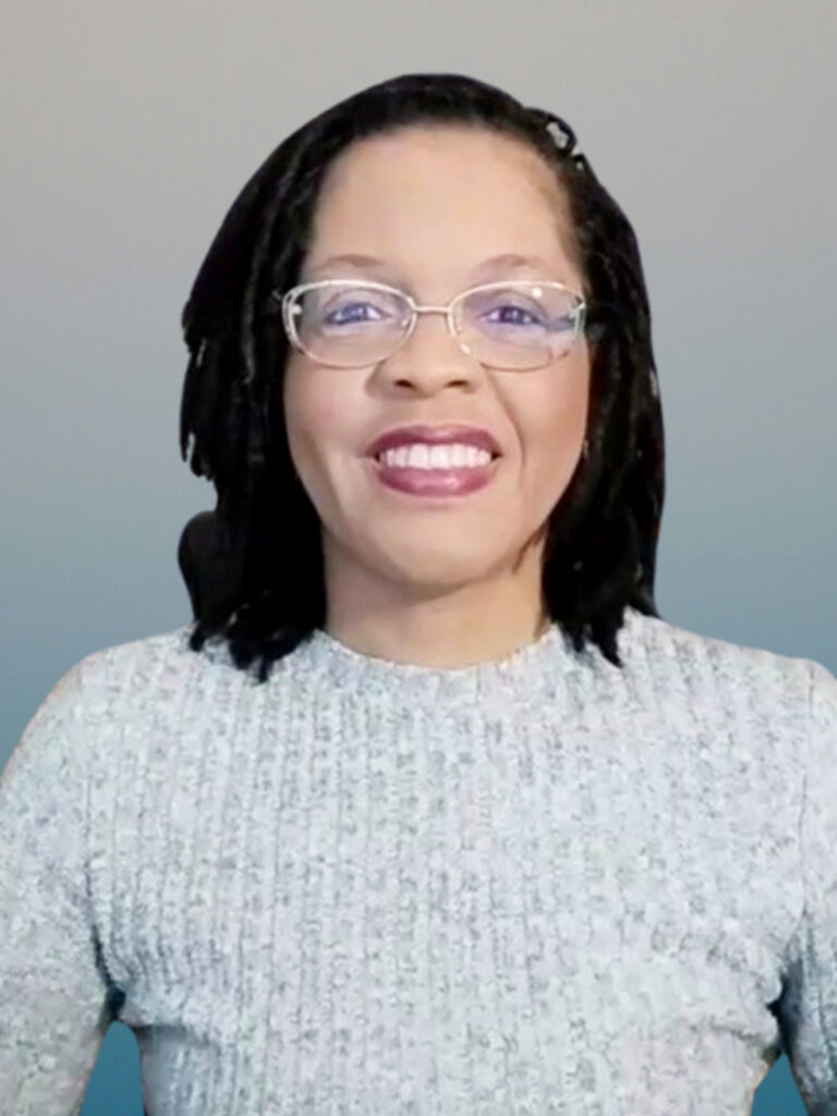 A picture of Toni Hogan wearing a light gray sweater. She is wearing glasses, with a microloc hairstyle.
